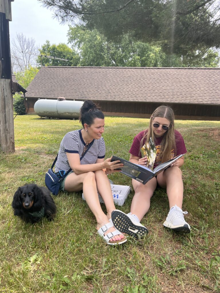 2 women looking at a book sitting on the grass with a dog nearby